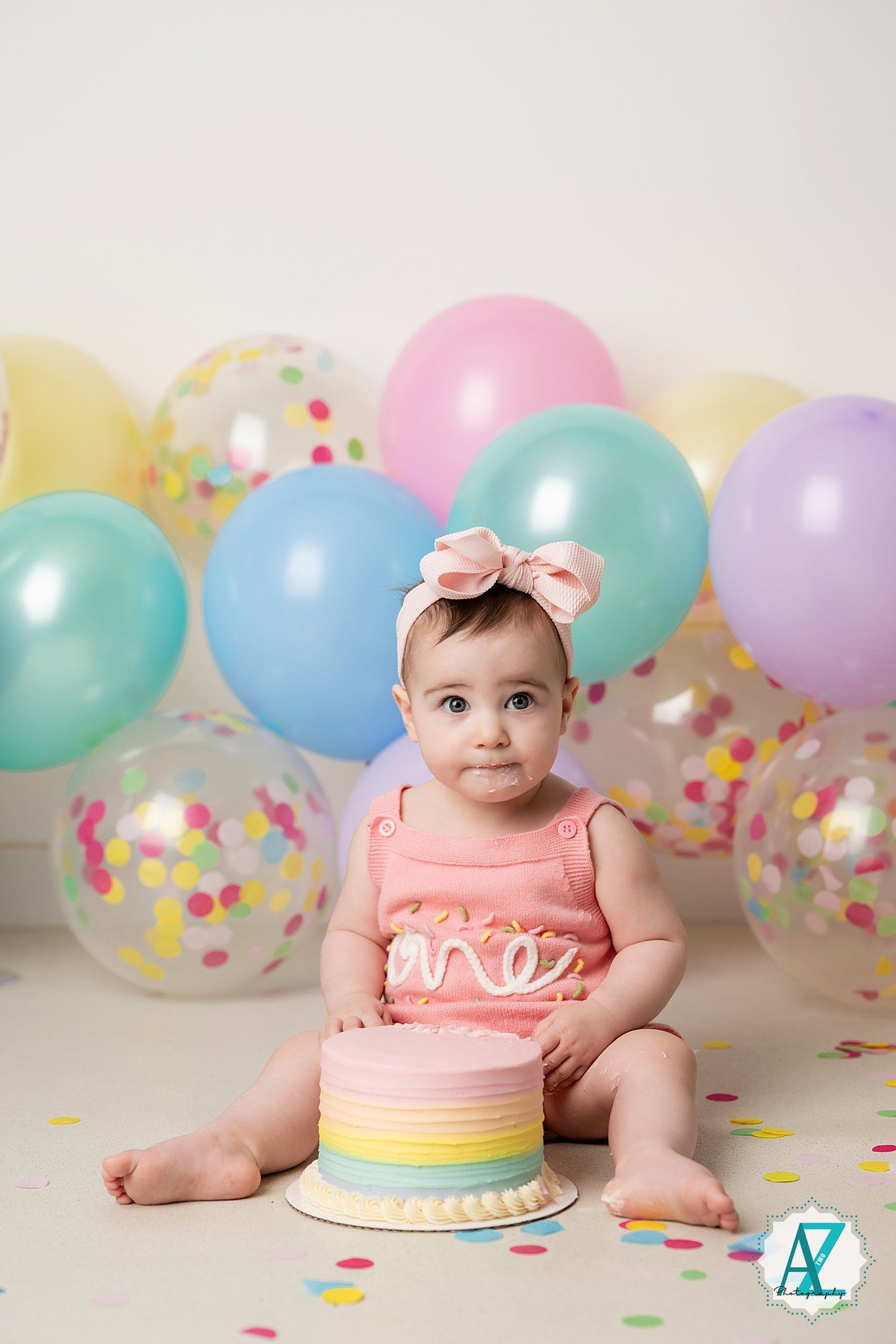 Baby in pink outfit with bow sitting by pastel rainbow cake surrounded by colorful balloons.