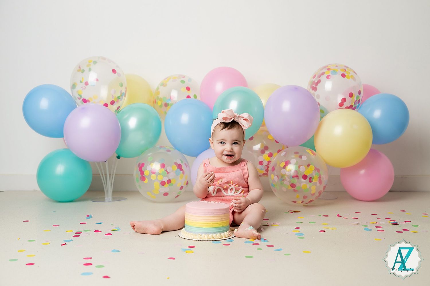 Festive first birthday setup with pastel balloons, confetti, and rainbow cake on white backdrop.