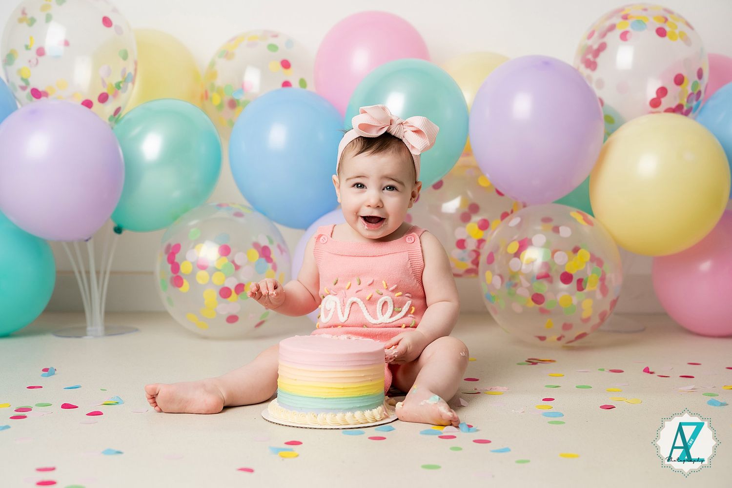 Baby laughing during cake smash photo session with colorful balloons and confetti background.