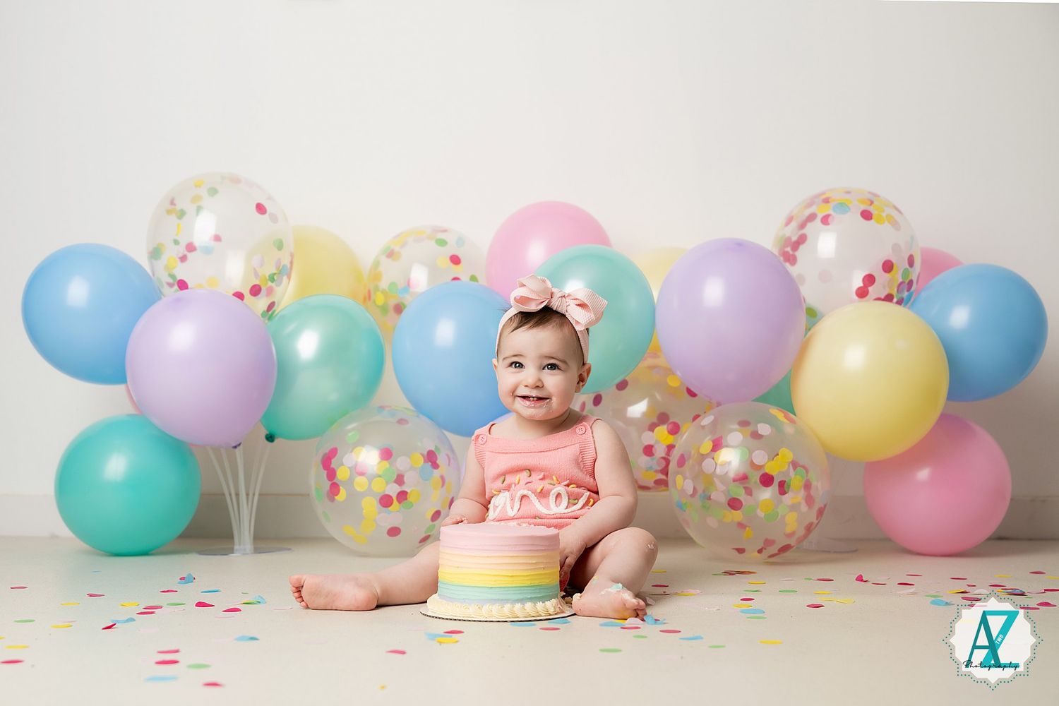 Baby in pink romper surrounded by pastel and confetti balloons with rainbow birthday cake.