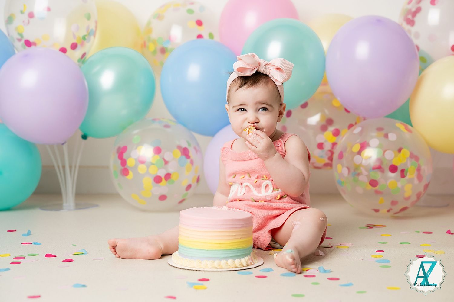 Baby in pink outfit reaching for pastel rainbow cake with colorful balloon backdrop.
