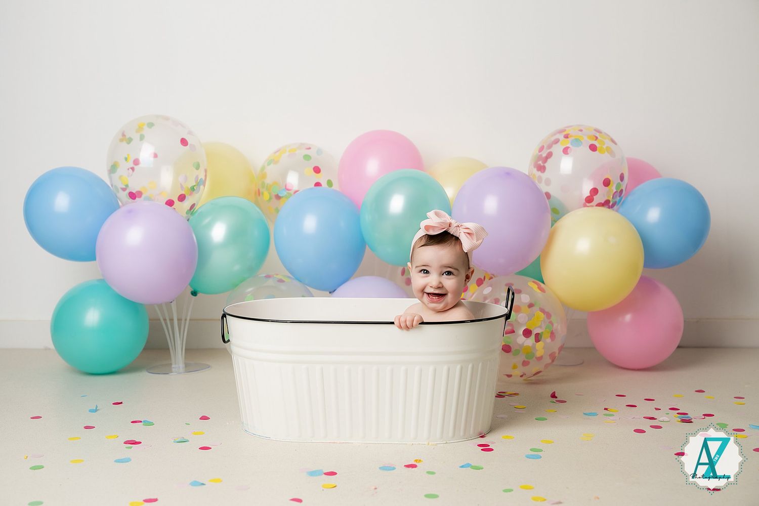 Baby sits in white washtub with colorful pastel balloons and confetti on light background for birthday celebration.