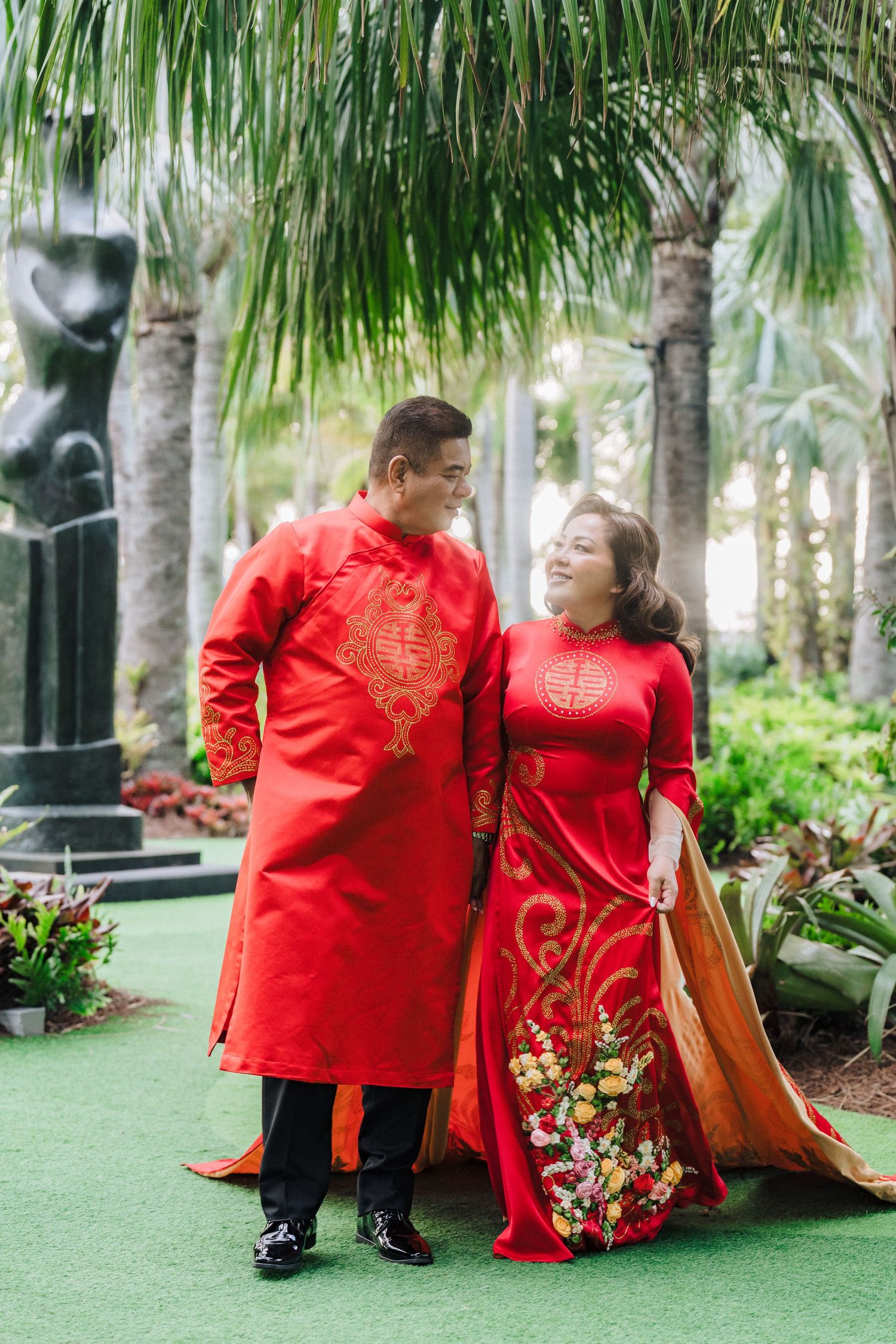 A couple wearing traditional red Vietnamese ao dai outfits smiles together in a tropical garden setting with palm trees. Sunset Bay Weddings