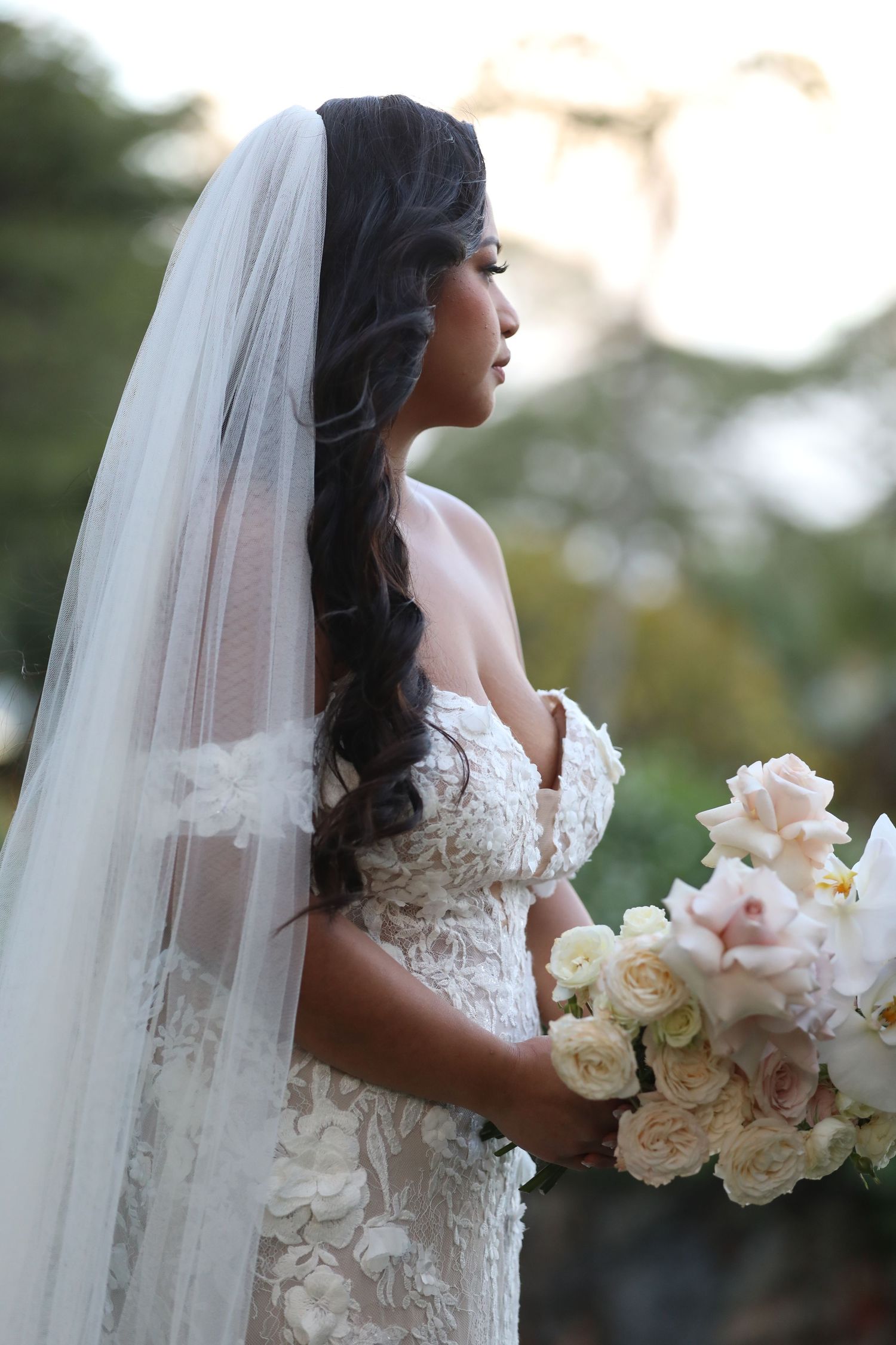 A side profile view of a bride wearing an elegant lace wedding dress and long veil while holding a soft pink bouquet.