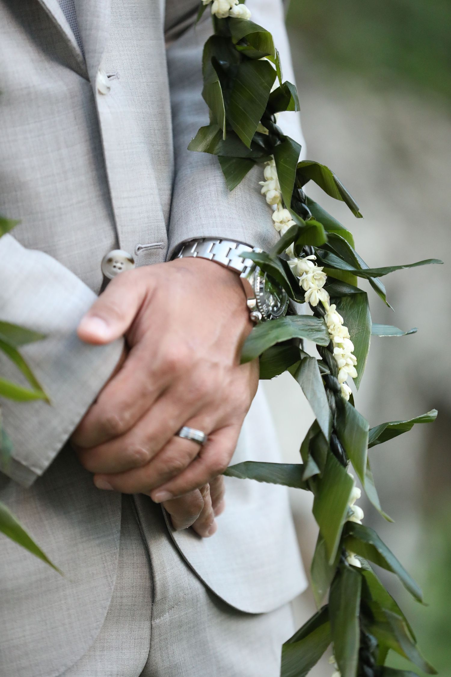 A hand wearing a metal watch and ring against a light gray suit sleeve with delicate white flowers and greenery.