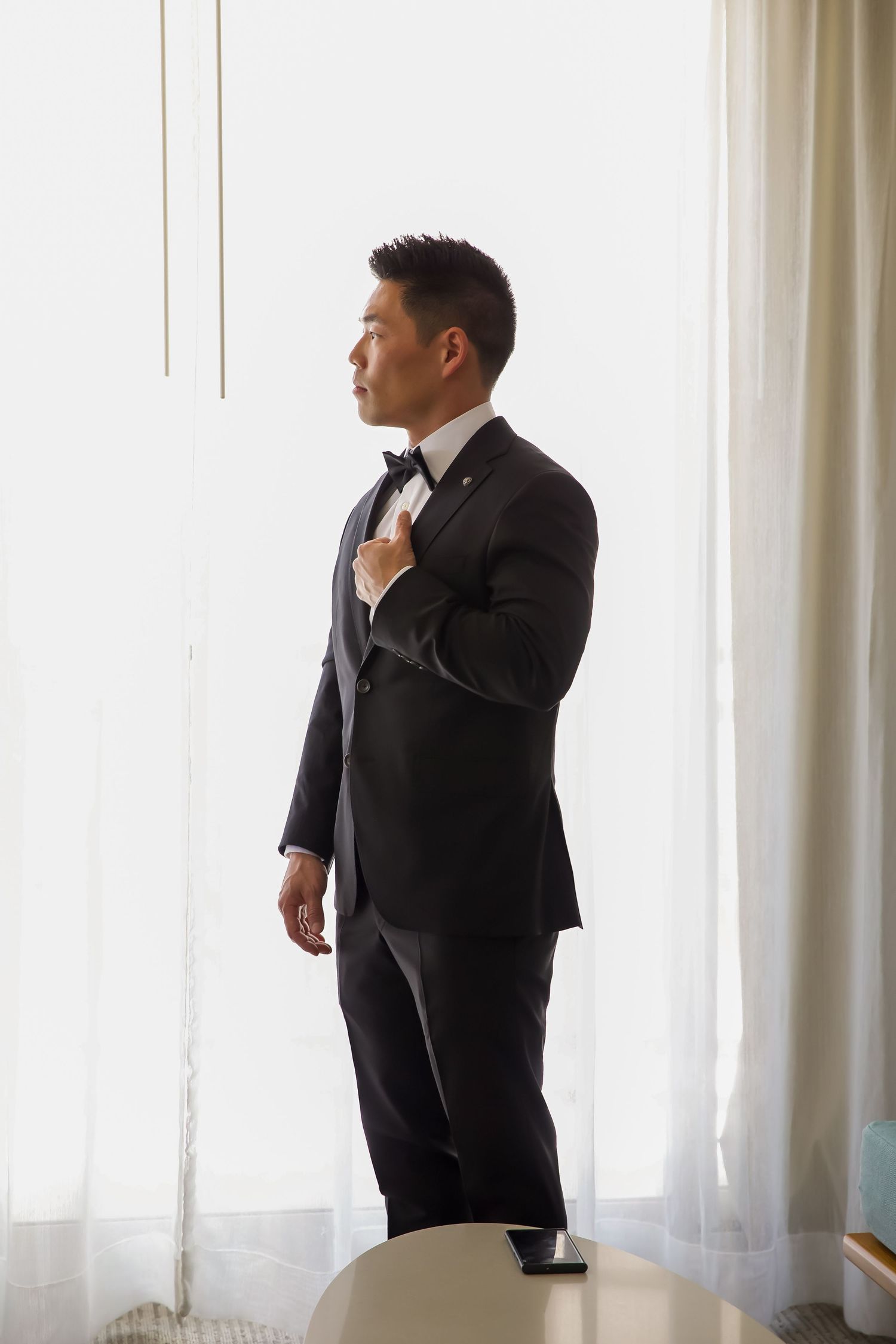 A person wearing a black suit and tie adjusts their bowtie while standing near white curtains in a bright room.