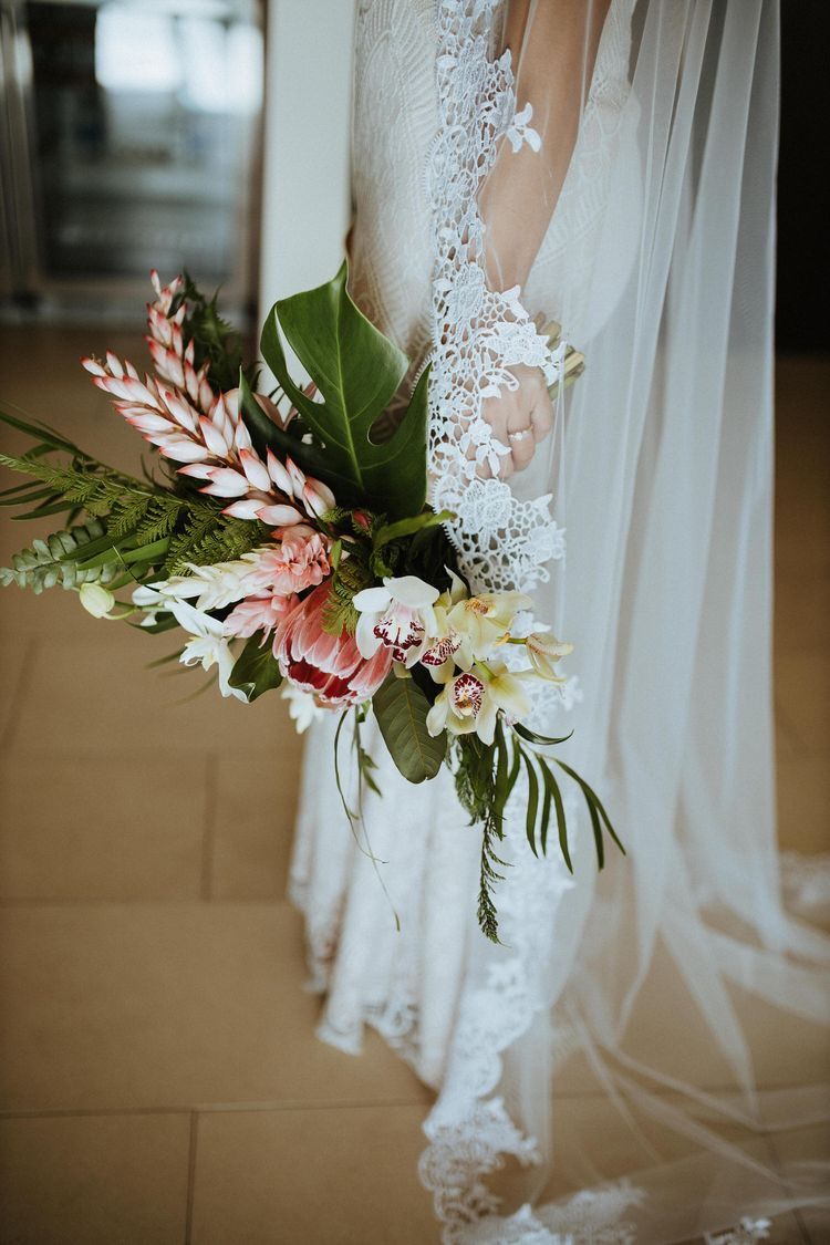 A romantic bridal bouquet with pink protea flowers and tropical greenery against delicate white lace wedding veil details.