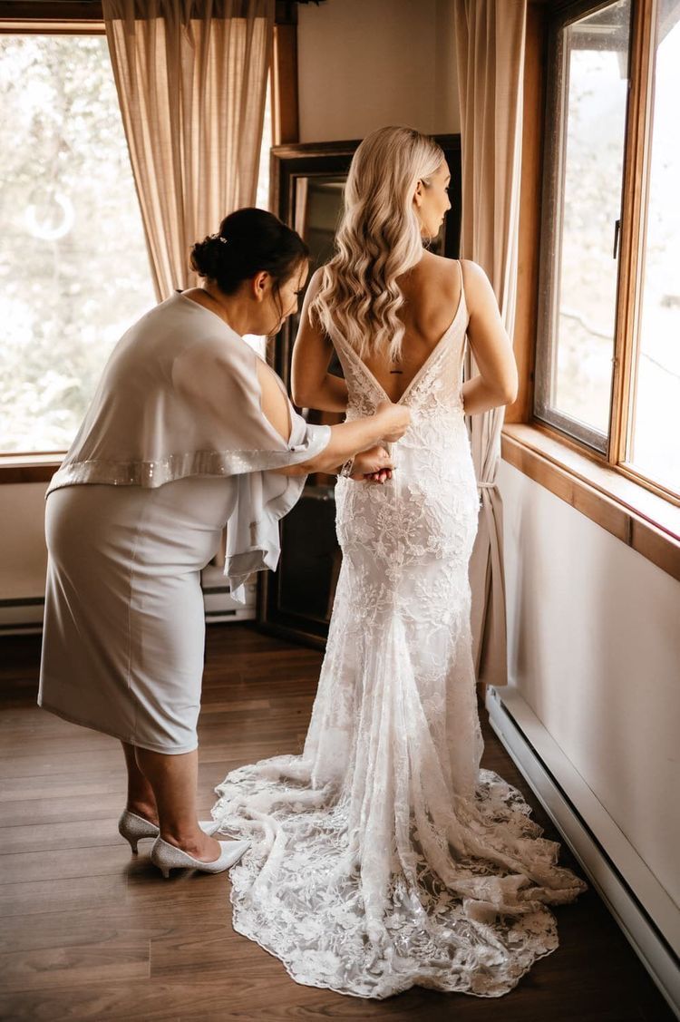 A woman helps button up an elegant lace wedding dress with a long train in a sunlit room.