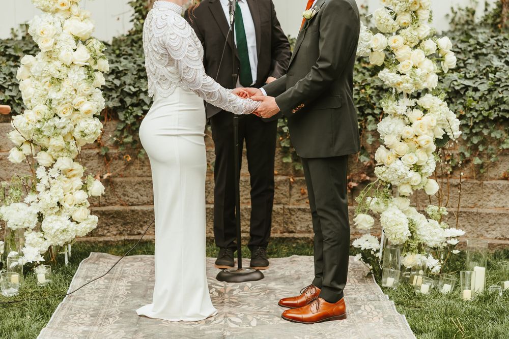 A couple holding hands during an outdoor wedding ceremony surrounded by white floral arrangements and a brick wall backdrop.