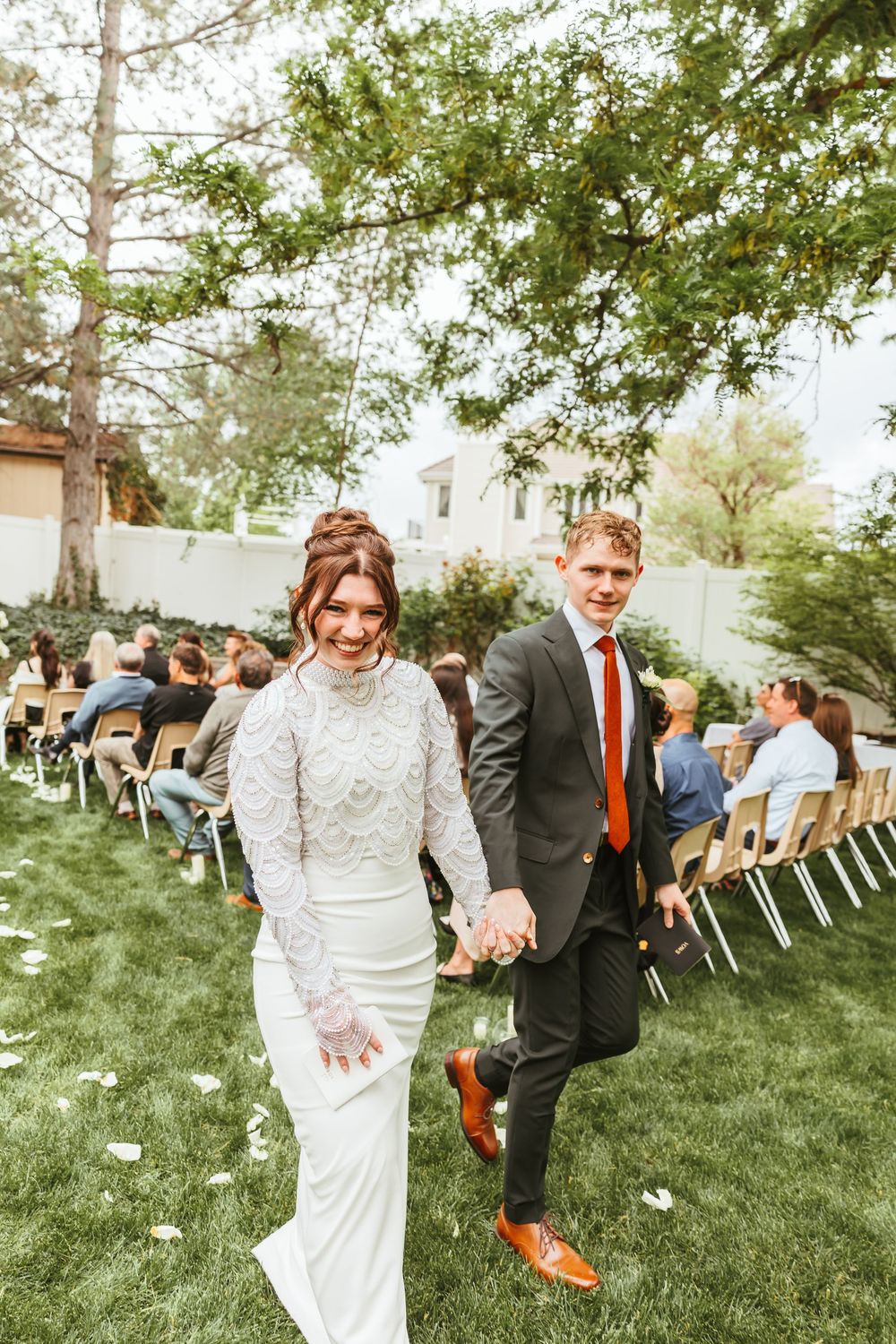 Couple exits outdoor wedding ceremony on lush grass holding hands while guests sit in rows of white chairs.
