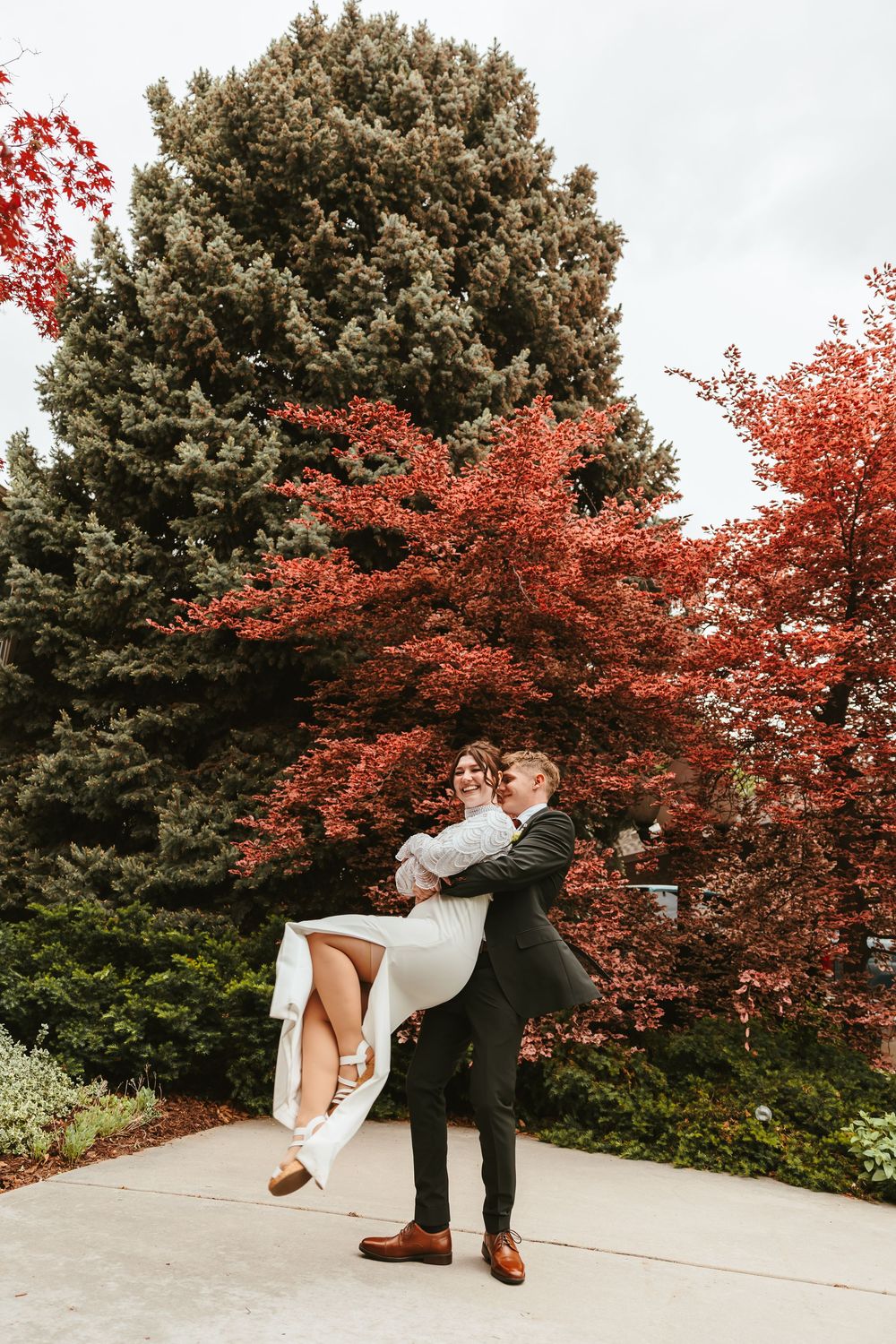 A couple embraces on a walkway surrounded by vibrant red fall foliage and a large evergreen tree.