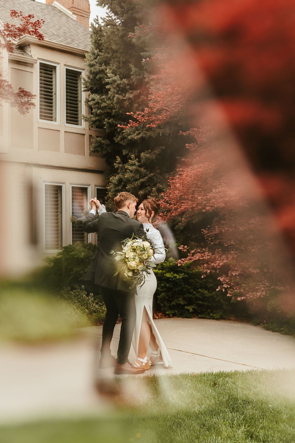 A romantic wedding portrait through red autumn foliage against a white house exterior.