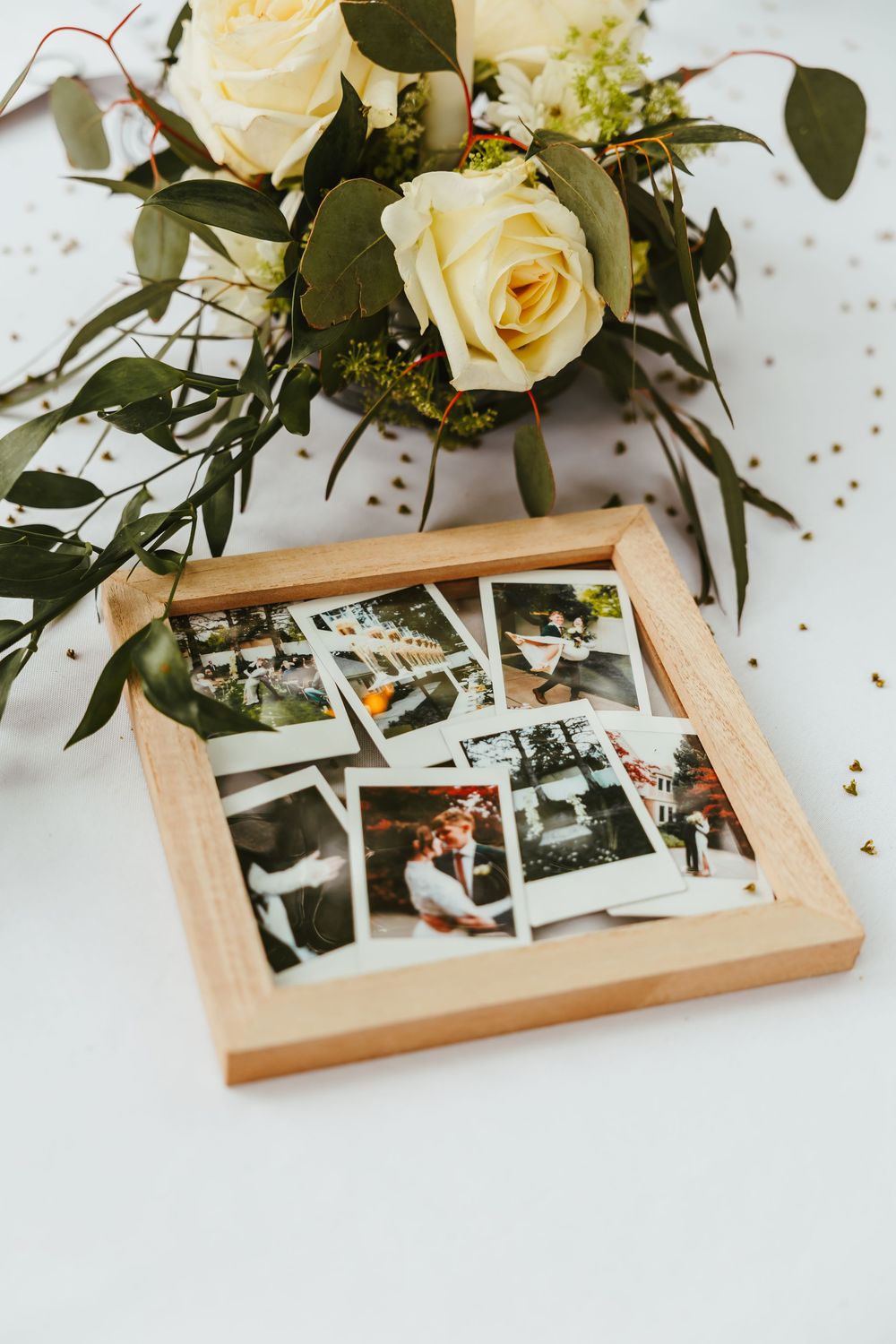 Cream roses and greenery arranged above wooden frame containing vintage photographs on white surface.