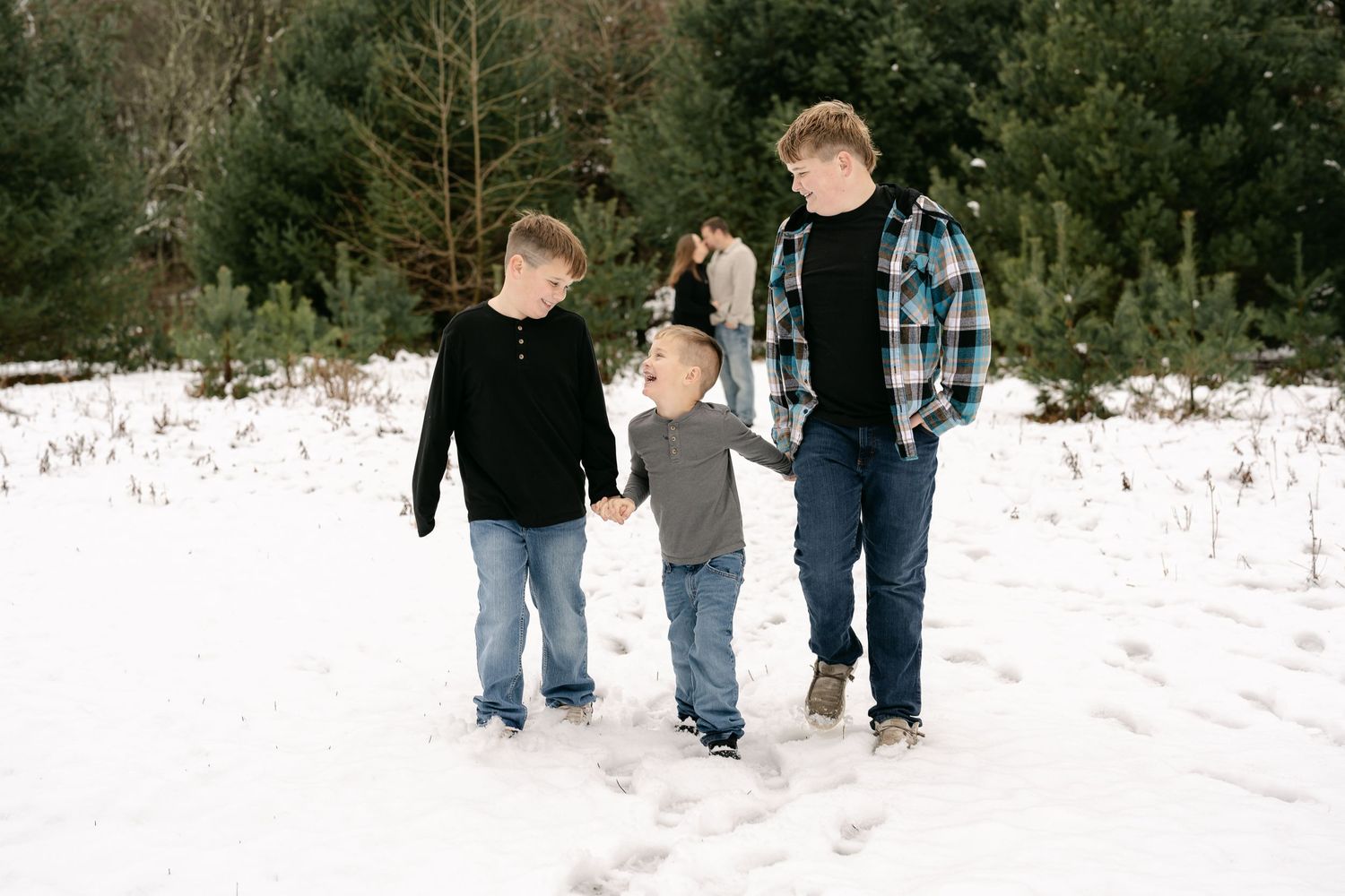 Three children walking together through snow-covered ground with evergreen trees in background.