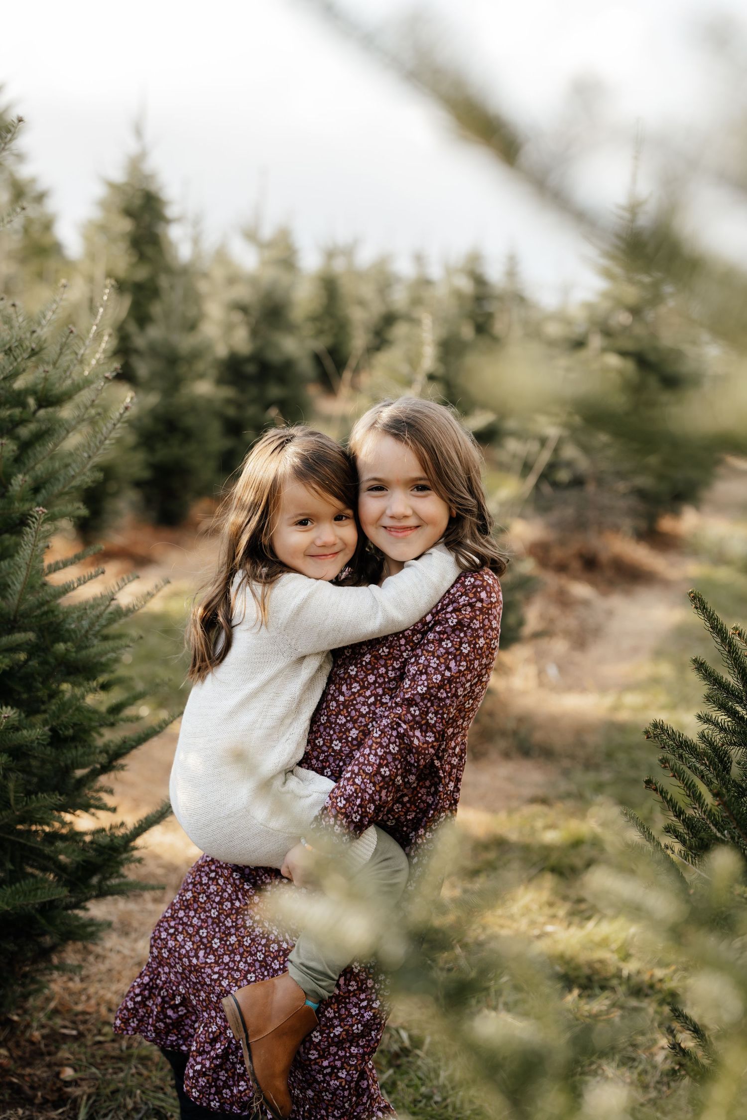Two children embrace while surrounded by evergreen trees on a cloudy day.