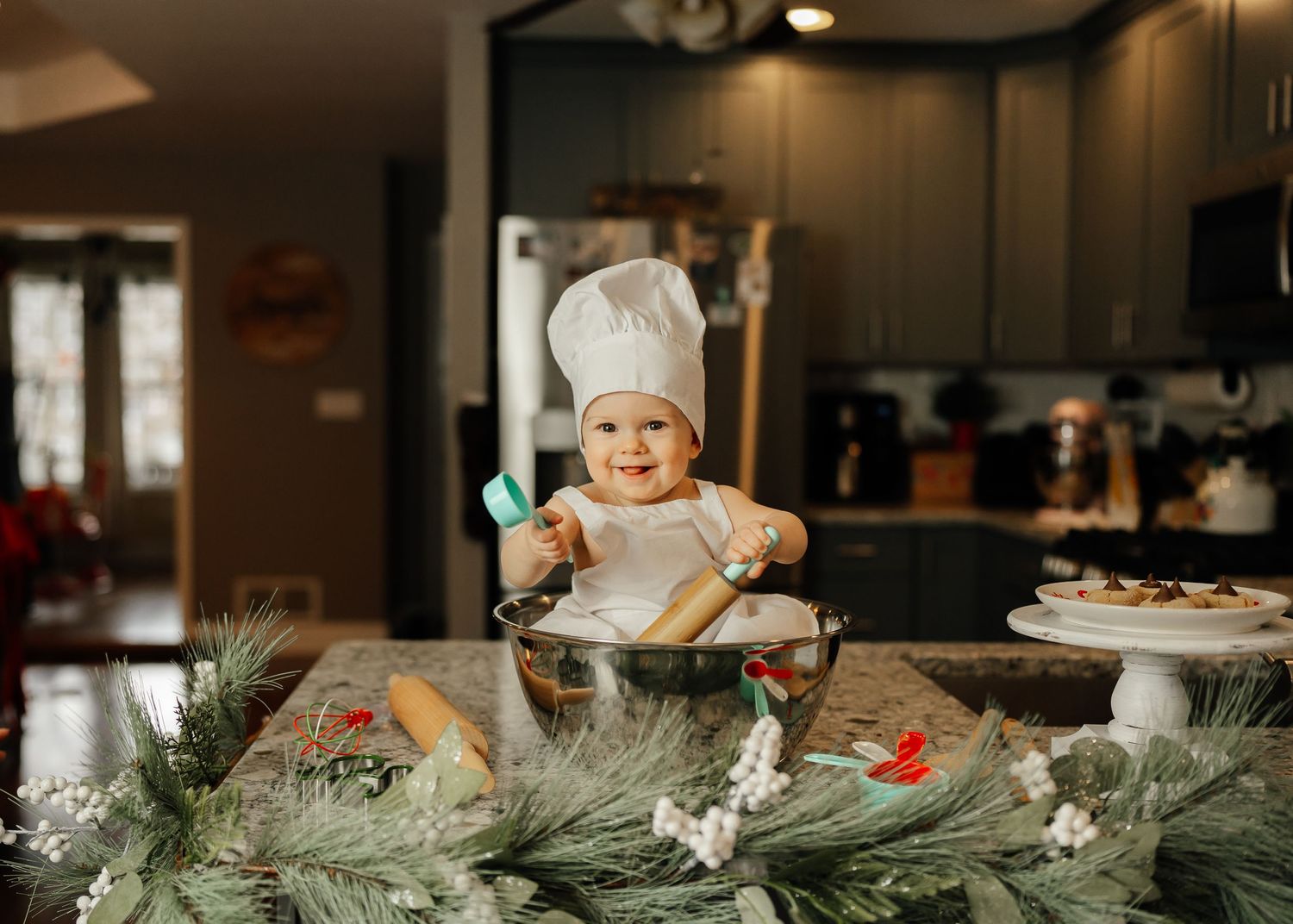 A baby in chef hat sits among Christmas decorations in kitchen.