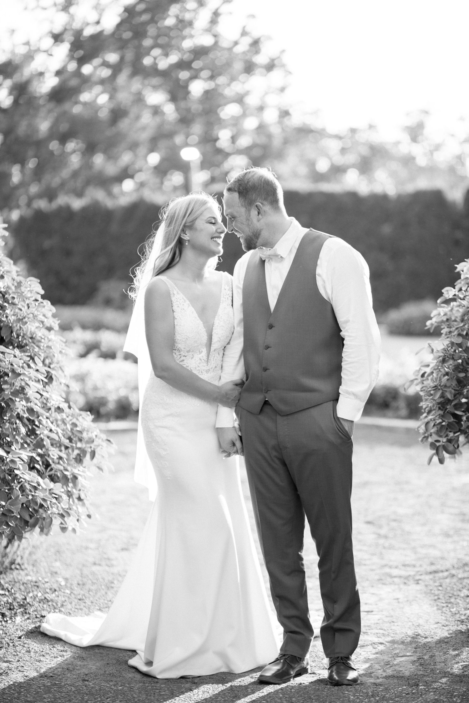 Black and white wedding photo of a couple standing together outdoors on a garden path surrounded by foliage.