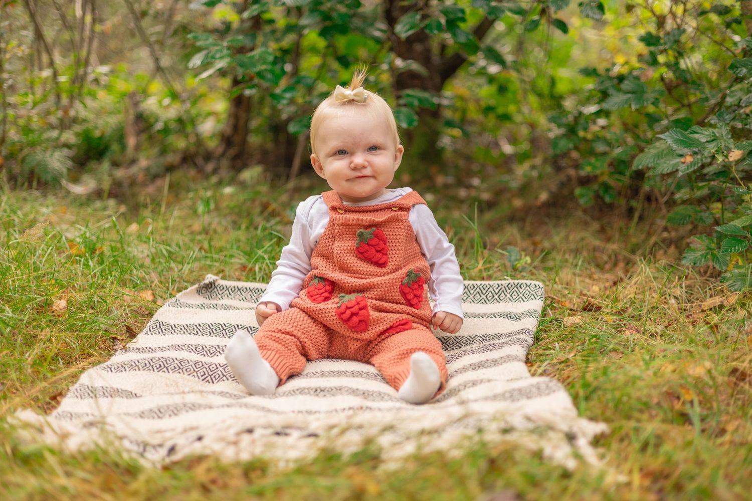 Baby in orange overalls sits on striped blanket in woodland setting.