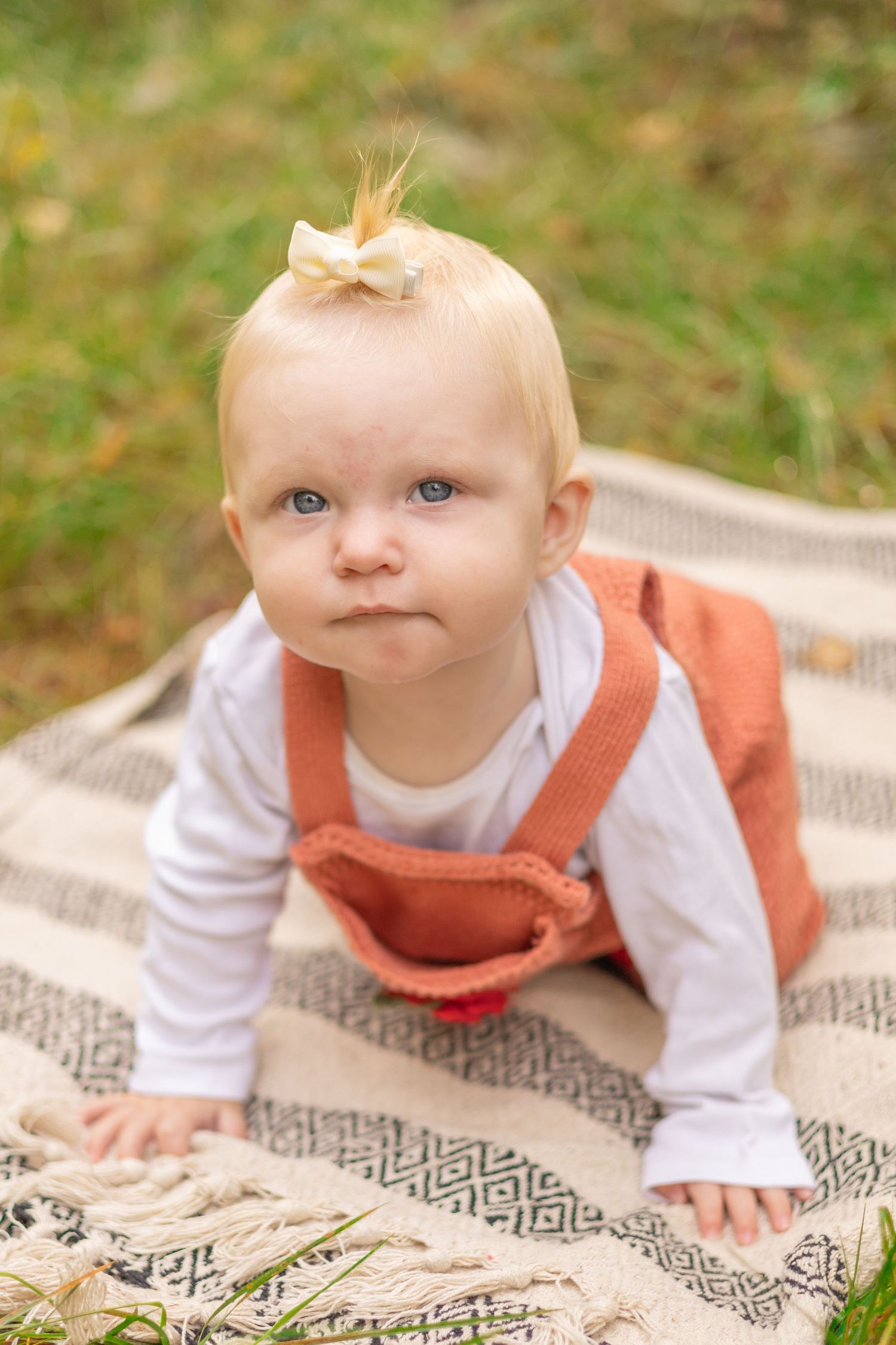 Baby in orange overalls crawls on patterned blanket outdoors with small bow in hair.