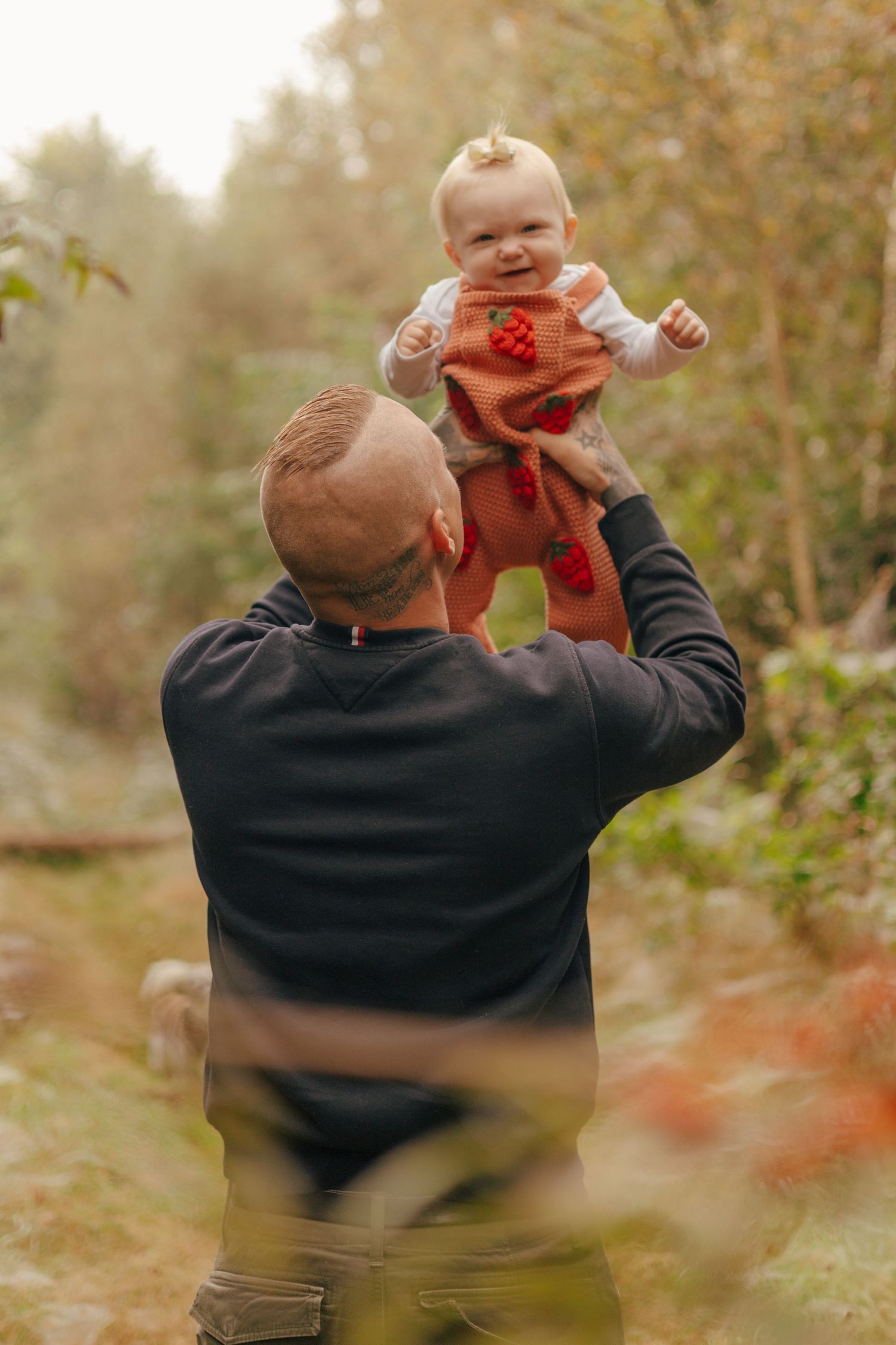 Adult lifts small child wearing orange outfit against autumn forest backdrop.