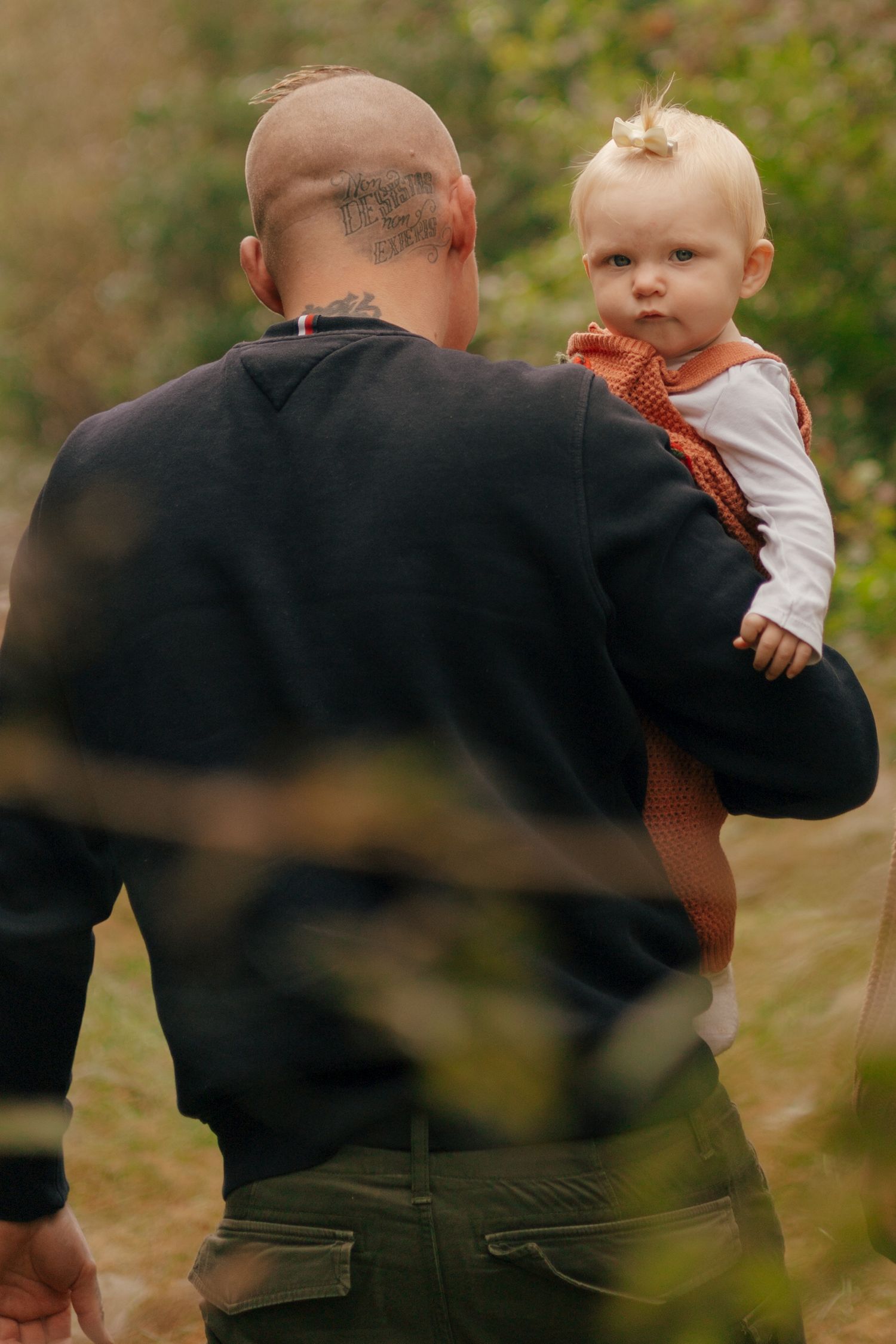Adult holds small child wearing orange outfit while standing in wooded area.
