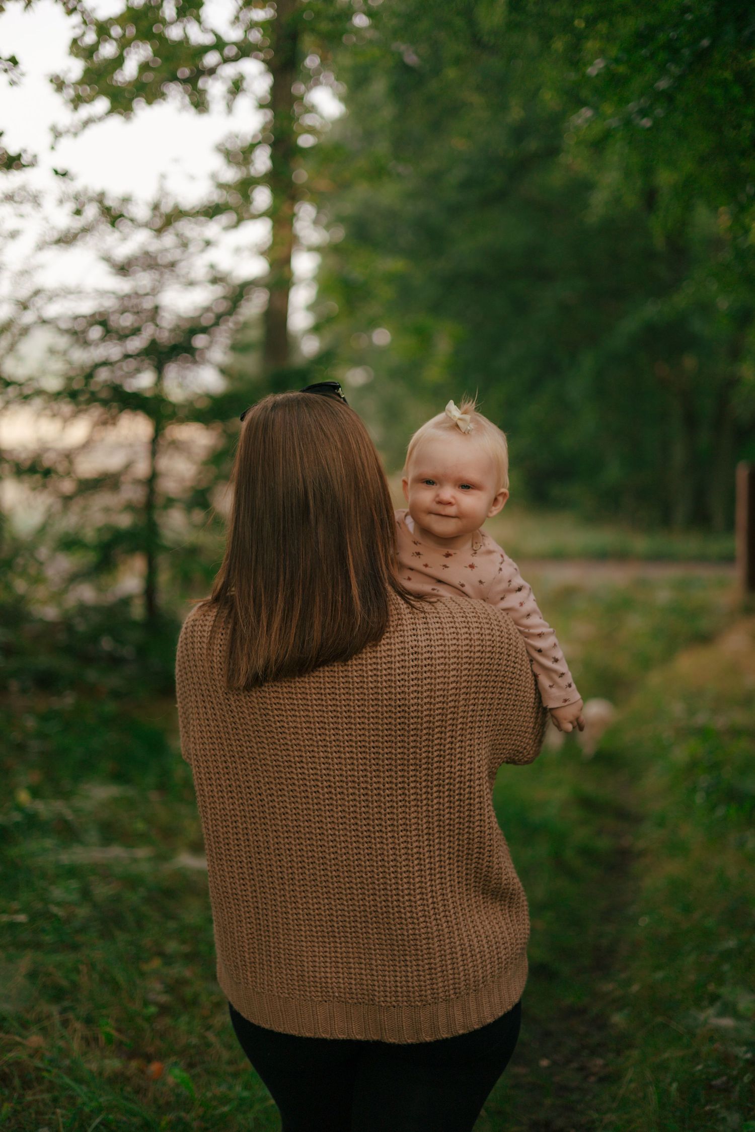 Person in brown sweater holds baby while standing in wooded path.