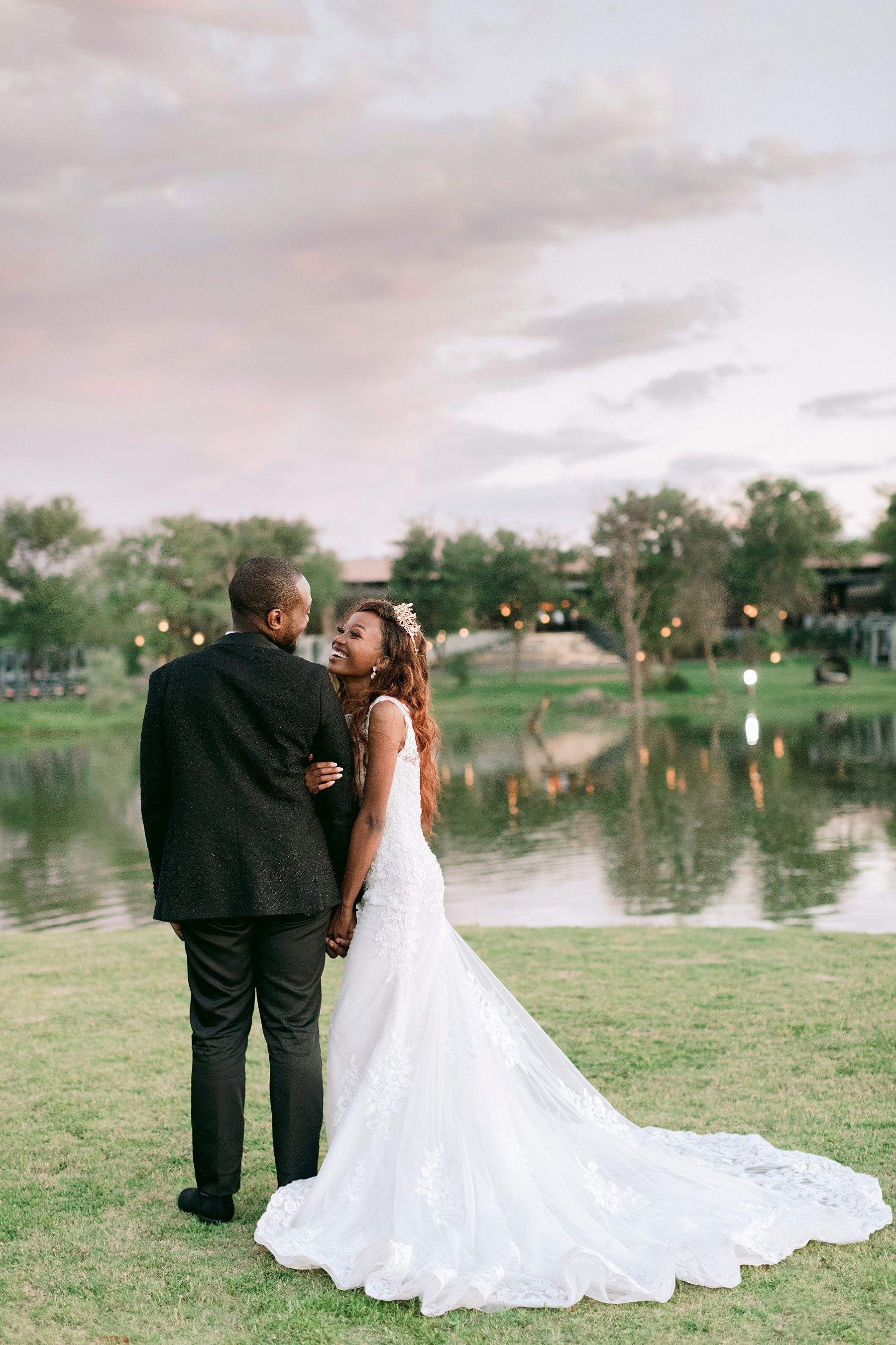 Bride and groom stand by a serene lake at sunset, bride\u2019s lace train flowing across green grass.