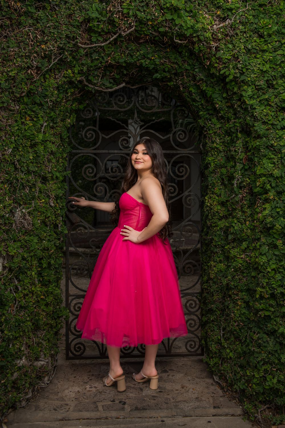 A hot pink formal dress creates contrast against an ornate iron gate covered in ivy.