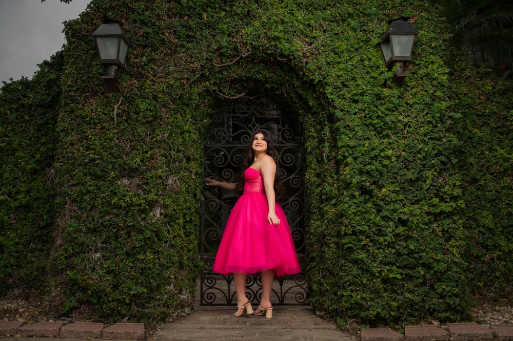 A bright fuchsia tea-length dress stands elegantly framed by an ivy-covered archway and lanterns.