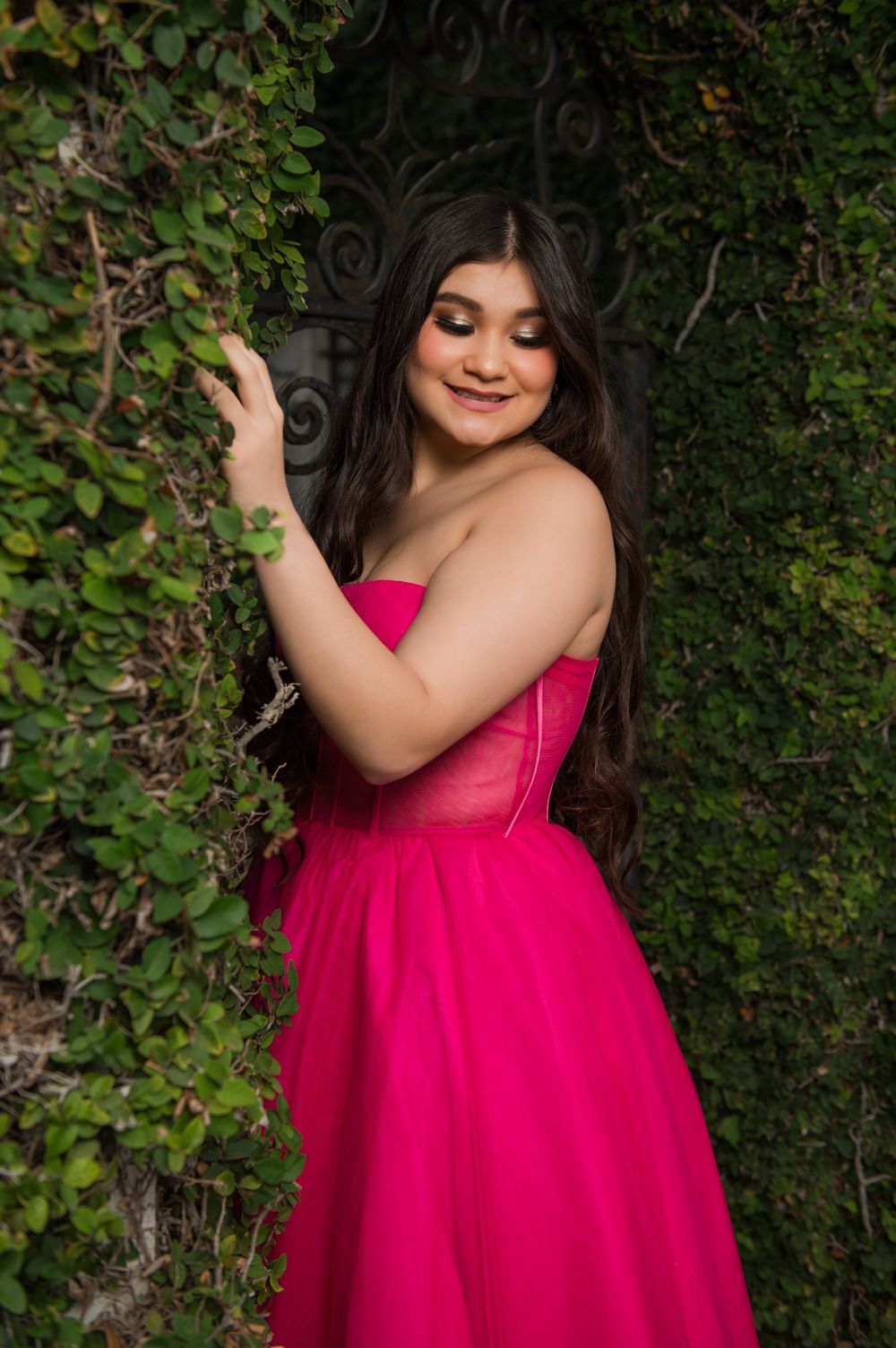 A bright pink strapless gown creates a striking contrast against a wall of green foliage.