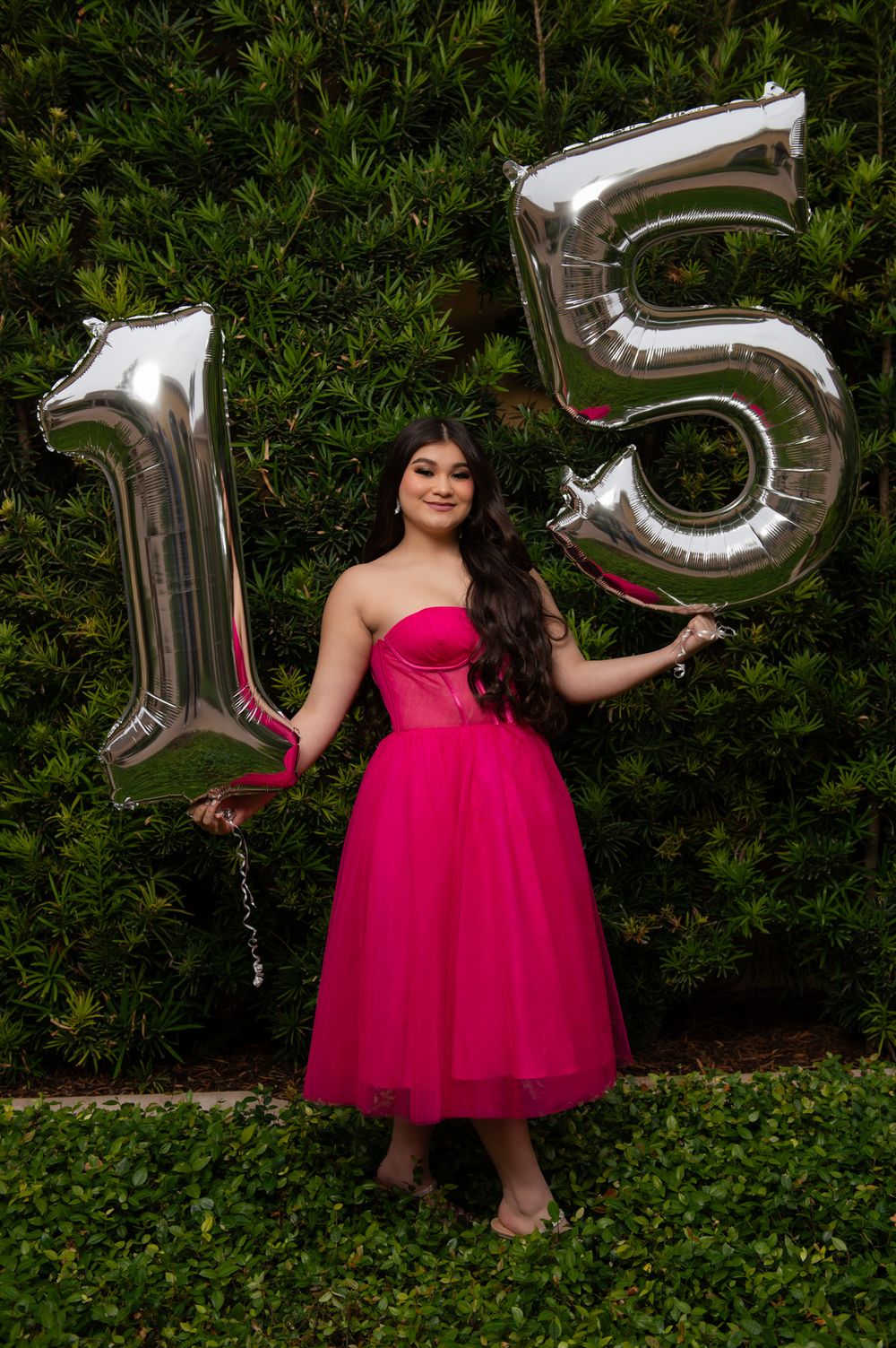 Festive quinceañera celebration with silver number balloons and a vibrant magenta tulle dress against greenery.