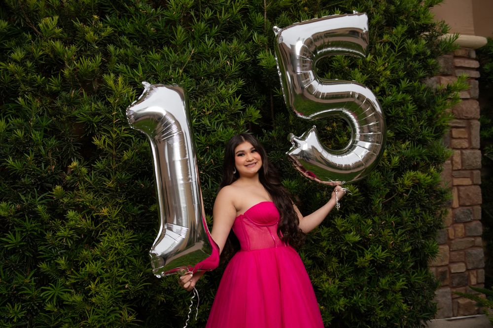 Silver number balloons form '15' alongside a magenta ball gown against a lush green hedge backdrop.