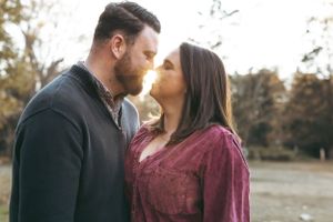 A couple shares an intimate moment outdoors during autumn, with the person in burgundy leaning towards their partner.