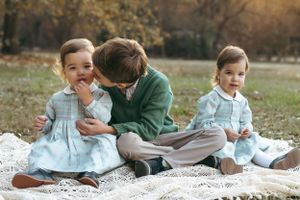 Three children in matching outfits sit on a blanket outdoors during autumn with golden sunlight in the background.