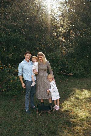 A family poses together outdoors in a wooded area with sunlight filtering through the trees in the background.