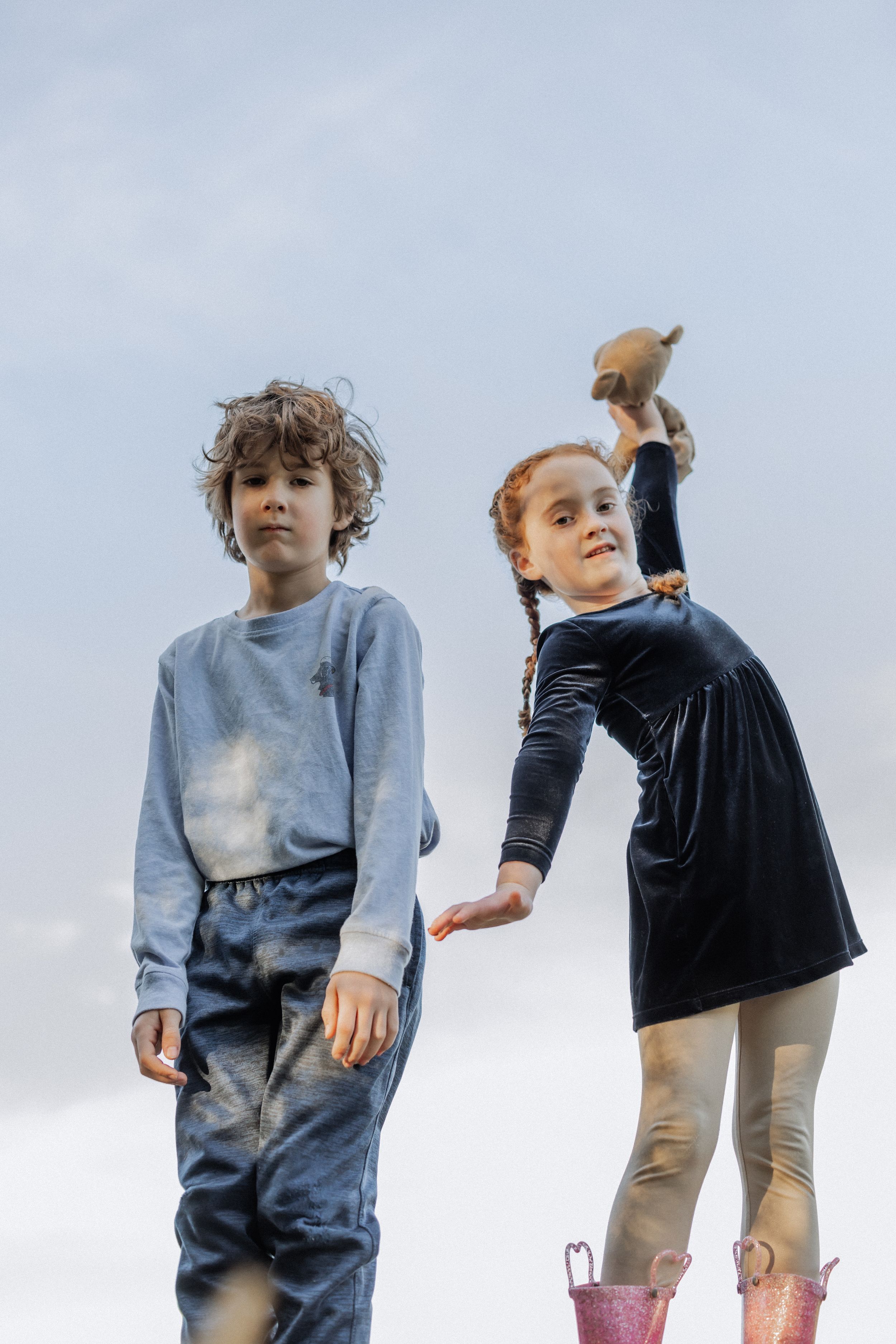 Two children stand against a blue sky, one in a dress holding something up high.