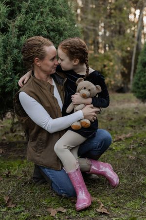 Parent and child share tender moment while holding teddy bear in forest setting.