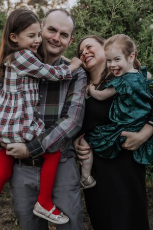 A joyful family portrait outdoors shows parents holding two children in coordinating holiday outfits.