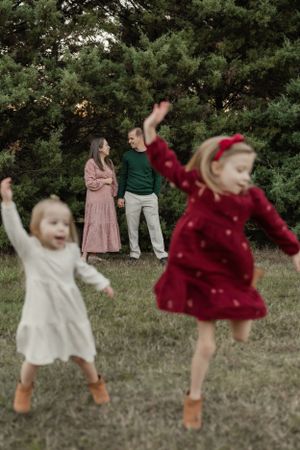 Children in dresses jumping joyfully in front of pine trees while adults watch.