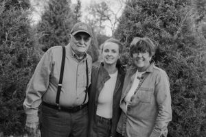 A black and white portrait of three people standing together in front of evergreen trees.