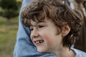A close-up portrait of a young child with curly hair smiling brightly outdoors.
