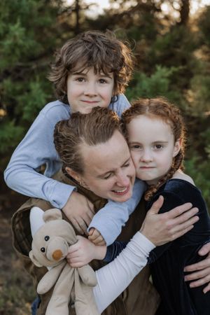 A joyful embrace between three people with a teddy bear outdoors among pine trees.