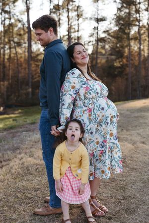 A family poses together outdoors with a pregnant woman in a floral dress, child in yellow, and tall figure in blue.