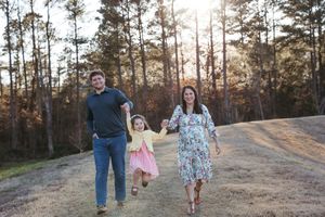 A family walks together on a dirt path during golden hour with sunlight filtering through pine trees in the background.