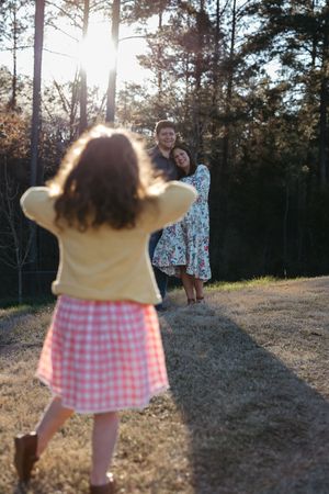 A young girl in a yellow top and pink gingham skirt approaches two figures on a sunlit forest path.