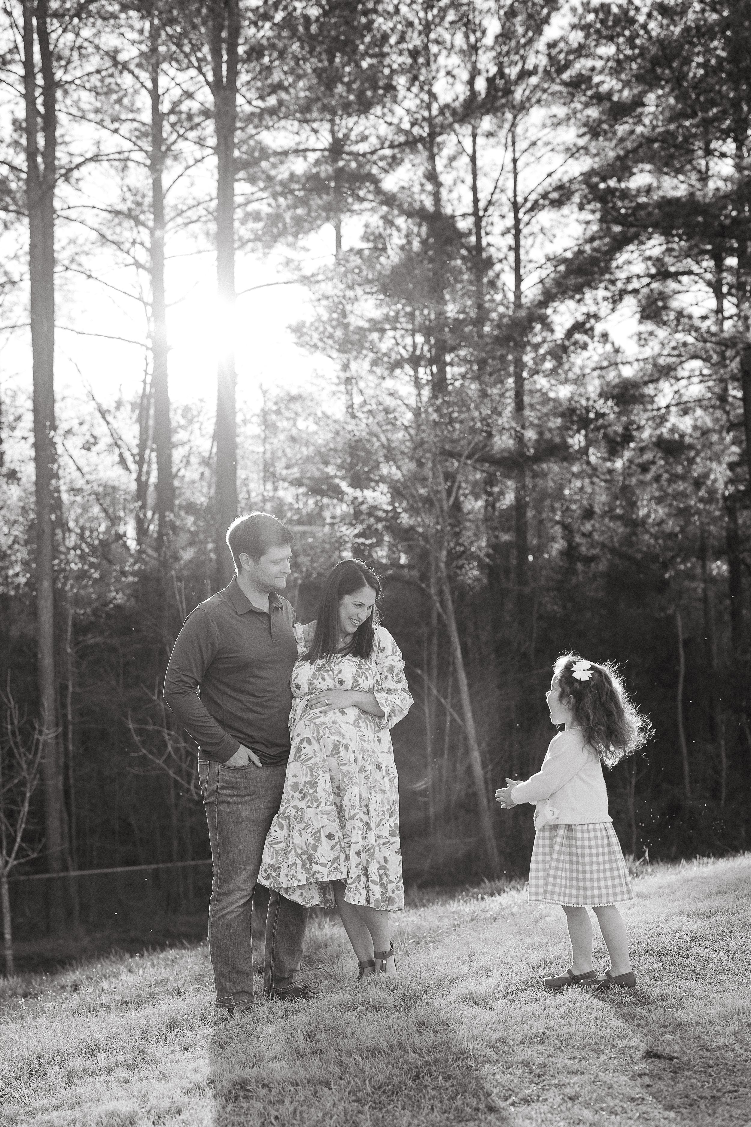 A black and white outdoor family portrait in a sunlit wooded area with trees creating a magical backlit effect.