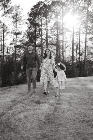 A family walks together on a grassy path through sunlit pine trees in black and white.