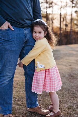 A child wearing a yellow cardigan and pink gingham dress stands next to someone in jeans on an autumn day.