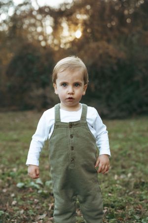 Small child stands in olive green overalls and white shirt outdoors at sunset with bokeh forest background.