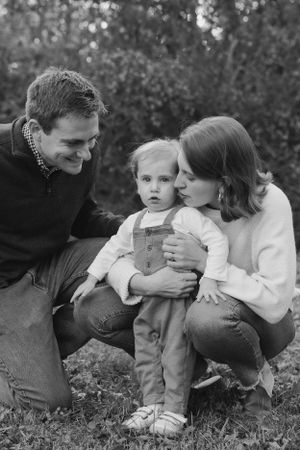 Black and white portrait of a family of three crouching together outdoors in casual clothing.